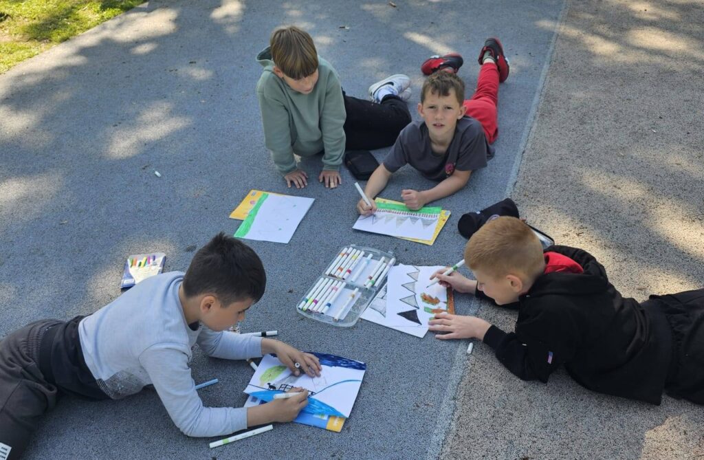 Uczniowie klasy III podczas plenerowej lekcji plastyki. Dzieci leżą na szkolnym boisku i tworzą kolorowe prace plastyczne inspirowane otaczającą przyrodą.