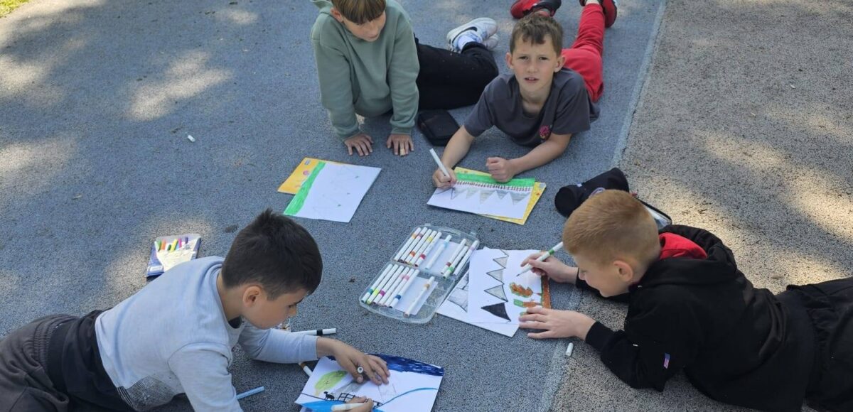 Uczniowie klasy III podczas plenerowej lekcji plastyki. Dzieci leżą na szkolnym boisku i tworzą kolorowe prace plastyczne inspirowane otaczającą przyrodą.