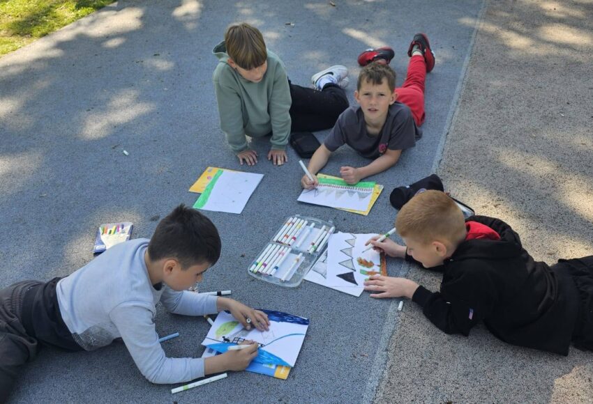 Uczniowie klasy III podczas plenerowej lekcji plastyki. Dzieci leżą na szkolnym boisku i tworzą kolorowe prace plastyczne inspirowane otaczającą przyrodą.