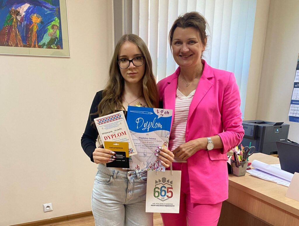 Uczennica klasy 8a Magdalena Imiołek prezentuje dyplom i nagrodę za zajęcie III miejsca w Miejskim Konkursie Ortograficznym „Mistrz Ortografii”. Obok stoi dyrektor szkoły.