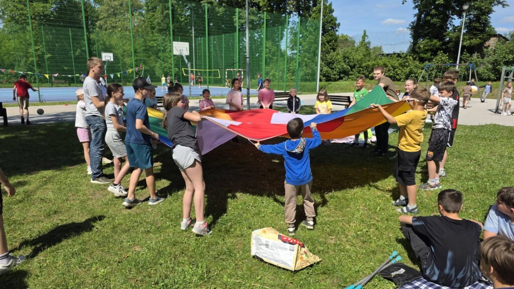 Uczniowie biorą udział w zabawie integracyjnej na szkolnym trawniku podczas obchodów Dnia Dziecka. Dzieci wspólnie trzymają kolorową chustę animacyjną, a w tle widoczne jest boisko szkolne oraz pozostali uczestnicy wydarzenia korzystający z przygotowanych atrakcji na świeżym powietrzu.
