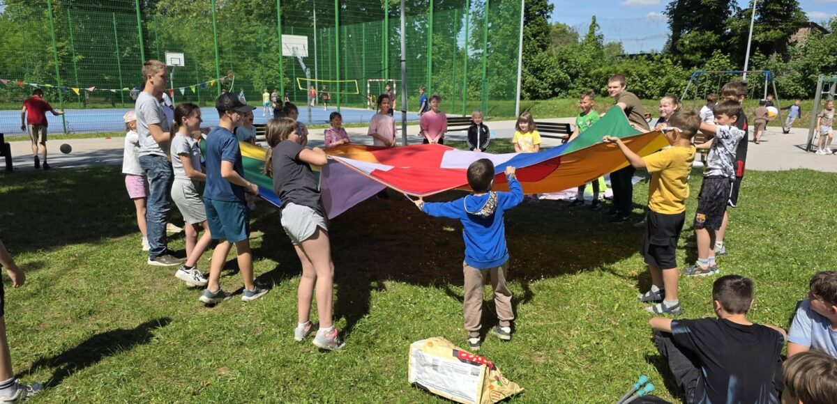 Uczniowie biorą udział w zabawie integracyjnej na szkolnym trawniku podczas obchodów Dnia Dziecka. Dzieci wspólnie trzymają kolorową chustę animacyjną, a w tle widoczne jest boisko szkolne oraz pozostali uczestnicy wydarzenia korzystający z przygotowanych atrakcji na świeżym powietrzu.