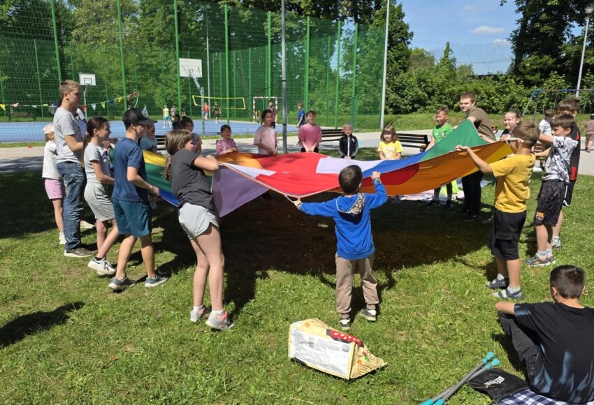 Uczniowie biorą udział w zabawie integracyjnej na szkolnym trawniku podczas obchodów Dnia Dziecka. Dzieci wspólnie trzymają kolorową chustę animacyjną, a w tle widoczne jest boisko szkolne oraz pozostali uczestnicy wydarzenia korzystający z przygotowanych atrakcji na świeżym powietrzu.