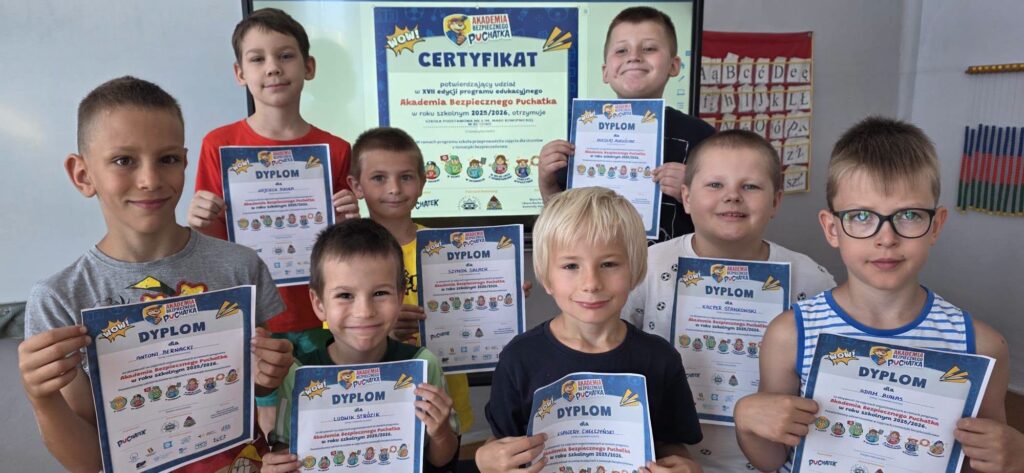 Uczniowie klasy pierwszej prezentują dyplomy otrzymane po zakończeniu programu „Akademia Bezpiecznego Puchatka”, a w tle widoczny jest certyfikat przyznany szkole.