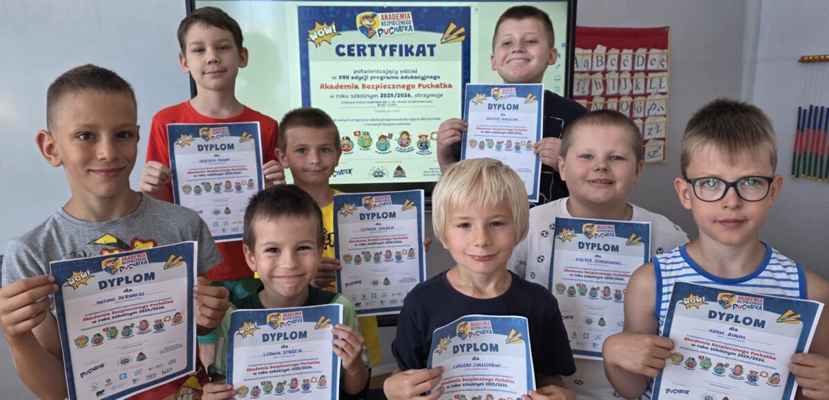 Uczniowie klasy pierwszej prezentują dyplomy otrzymane po zakończeniu programu „Akademia Bezpiecznego Puchatka”, a w tle widoczny jest certyfikat przyznany szkole.