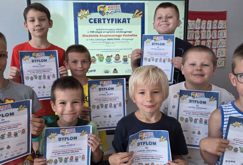 Uczniowie klasy pierwszej prezentują dyplomy otrzymane po zakończeniu programu „Akademia Bezpiecznego Puchatka”, a w tle widoczny jest certyfikat przyznany szkole.