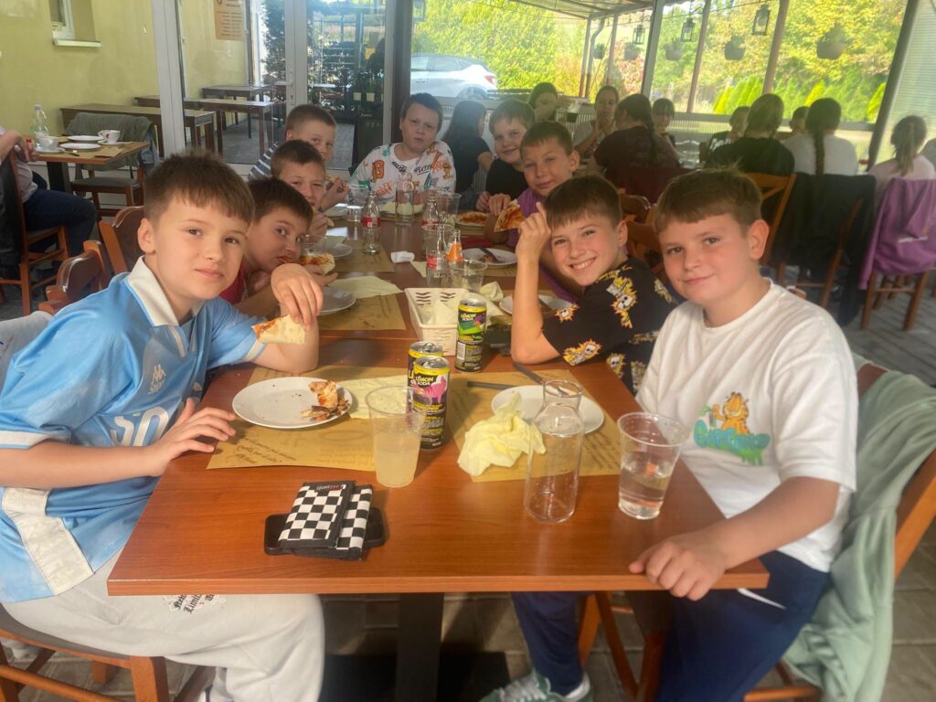 Uczniowie klas Va i VIa Szkoły Podstawowej nr 6 w Będzinie podczas wspólnego wyjścia do Pizzerii Veneto z okazji Dnia Chłopaka. Chłopcy siedzą przy stole, jedzą pizzę i pozują do zdjęcia.