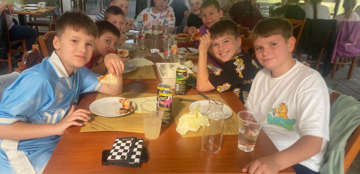 Uczniowie klas Va i VIa Szkoły Podstawowej nr 6 w Będzinie podczas wspólnego wyjścia do Pizzerii Veneto z okazji Dnia Chłopaka. Chłopcy siedzą przy stole, jedzą pizzę i pozują do zdjęcia.