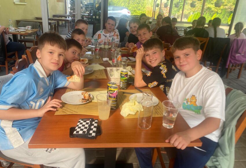 Uczniowie klas Va i VIa Szkoły Podstawowej nr 6 w Będzinie podczas wspólnego wyjścia do Pizzerii Veneto z okazji Dnia Chłopaka. Chłopcy siedzą przy stole, jedzą pizzę i pozują do zdjęcia.