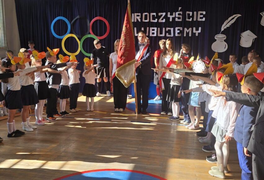 Uroczyste ślubowanie pierwszoklasistów Szkoły Podstawowej nr 6 w Będzinie. Uczniowie w strojach Kurczaczków i Kaczuszek, z wyciągniętymi rękami, składają ślubowanie w obecności pocztu sztandarowego podczas Podwórkowej Olimpiady.