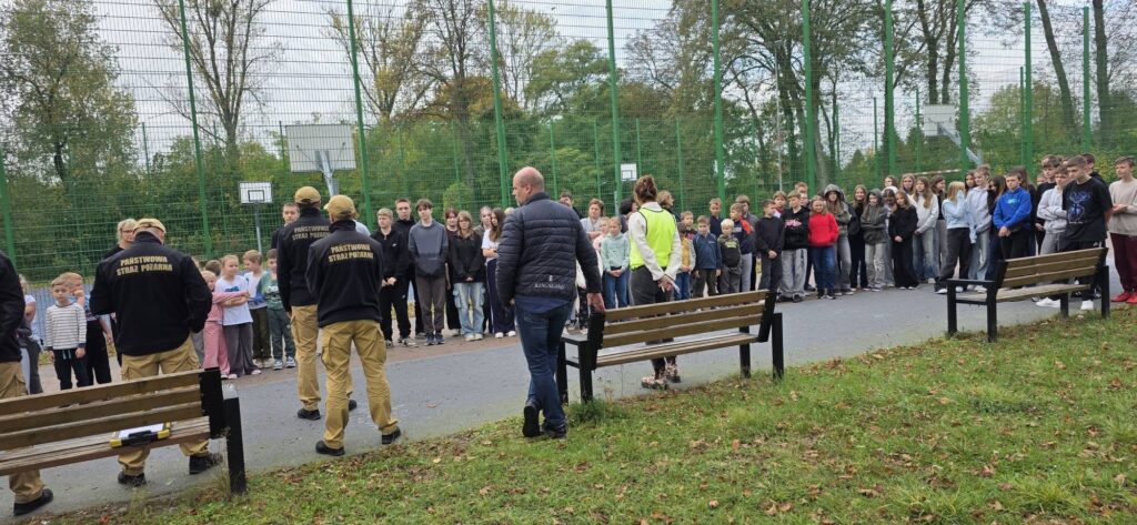 Uczniowie i nauczyciele zgromadzeni na boisku szkolnym podczas próbnej ewakuacji i spotkania z funkcjonariuszami Straży Pożarnej.