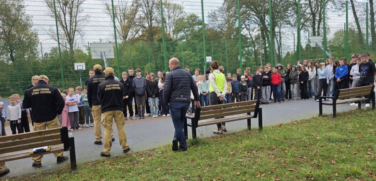 Uczniowie i nauczyciele zgromadzeni na boisku szkolnym podczas próbnej ewakuacji i spotkania z funkcjonariuszami Straży Pożarnej.