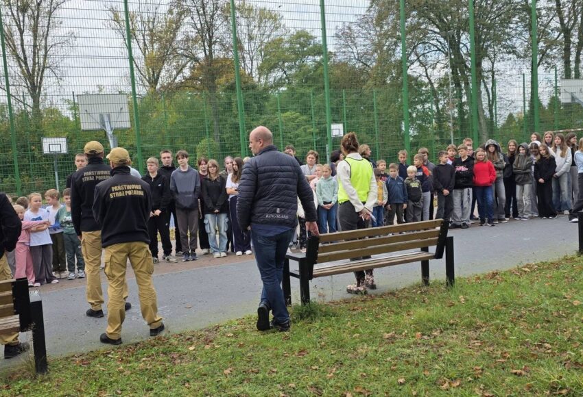 Uczniowie i nauczyciele zgromadzeni na boisku szkolnym podczas próbnej ewakuacji i spotkania z funkcjonariuszami Straży Pożarnej.