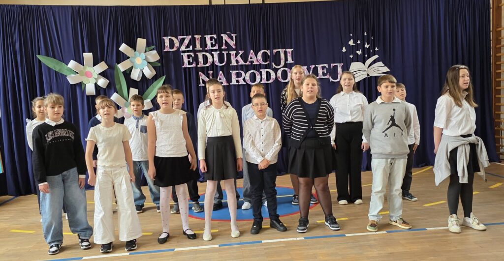 Grupa uczniów prezentuje występ wokalny podczas akademii z okazji Dnia Edukacji Narodowej. W tle granatowa kurtyna z napisem „Dzień Edukacji Narodowej”, ozdobiona kolorowymi kwiatami oraz dekoracją przedstawiającą otwartą książkę i motyle.