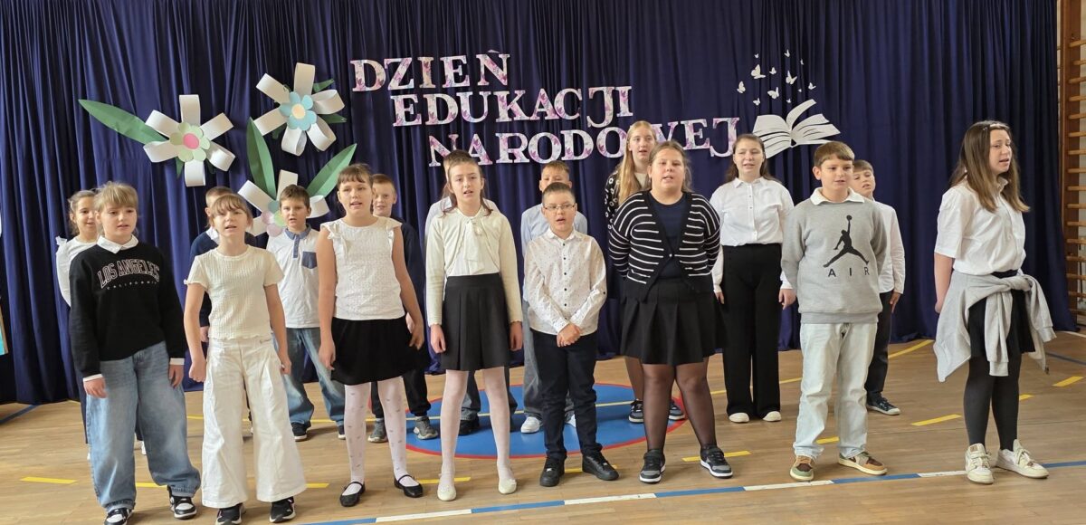 Grupa uczniów prezentuje występ wokalny podczas akademii z okazji Dnia Edukacji Narodowej. W tle granatowa kurtyna z napisem „Dzień Edukacji Narodowej”, ozdobiona kolorowymi kwiatami oraz dekoracją przedstawiającą otwartą książkę i motyle.