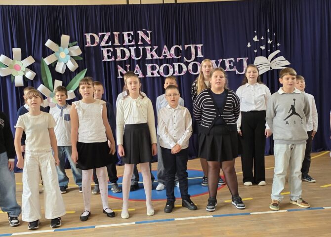 Grupa uczniów prezentuje występ wokalny podczas akademii z okazji Dnia Edukacji Narodowej. W tle granatowa kurtyna z napisem „Dzień Edukacji Narodowej”, ozdobiona kolorowymi kwiatami oraz dekoracją przedstawiającą otwartą książkę i motyle.