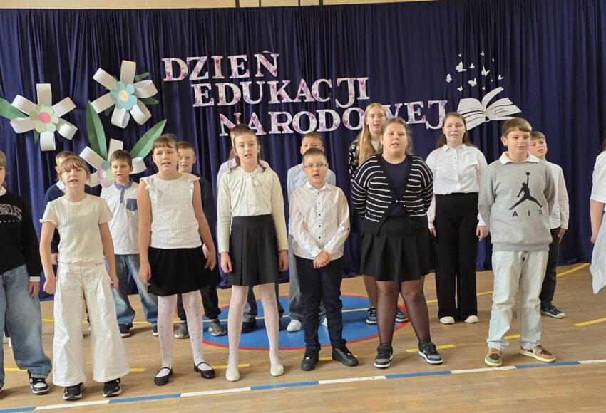 Grupa uczniów prezentuje występ wokalny podczas akademii z okazji Dnia Edukacji Narodowej. W tle granatowa kurtyna z napisem „Dzień Edukacji Narodowej”, ozdobiona kolorowymi kwiatami oraz dekoracją przedstawiającą otwartą książkę i motyle.