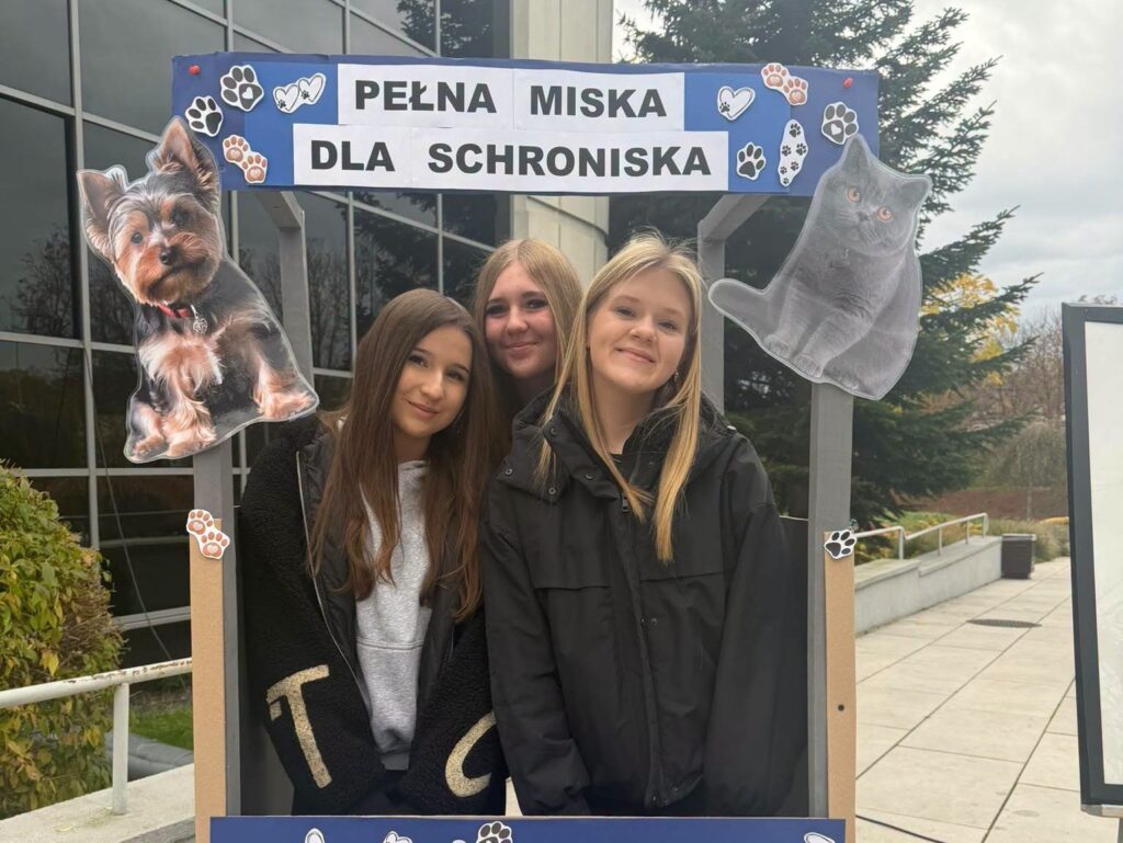 Trzy uczennice Szkoły Podstawowej nr 6 w Będzinie podczas XIV edycji akcji charytatywnej „Pełna Miska dla Schroniska”, przed dekoracyjną ramką z wizerunkami psa i kota.